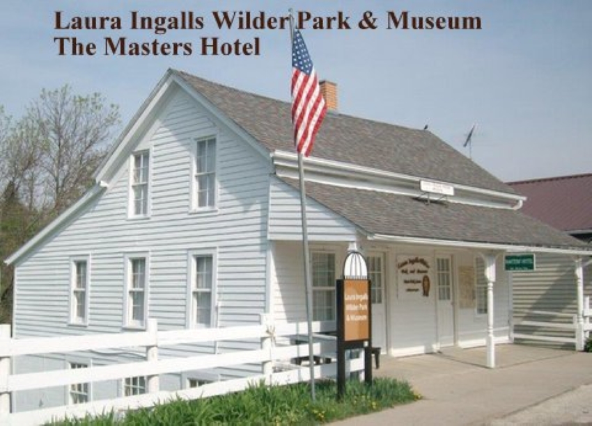 A white building with a gray roof and a roof over the sidewalk in front, supported on 3 slender pillars. The house has 2 stories, with another story partially visible down below street level on the side, where the ground slopes down. A flag on a flagpole in front of the building. Text above it: Laura Ingalls Wilder Park & Museum The Masters Hotel.