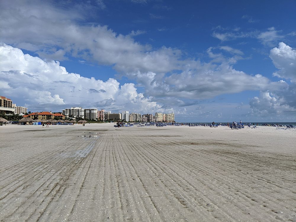 A view along the wide white-sand beach. In the foreground, the sand is flat and empty and looks like it's been raked. In the background, a cluster of tall buildings, presumably hotels and apartments. A bit of the ocean is visible on the right. White fluffy clouds and blue sky.