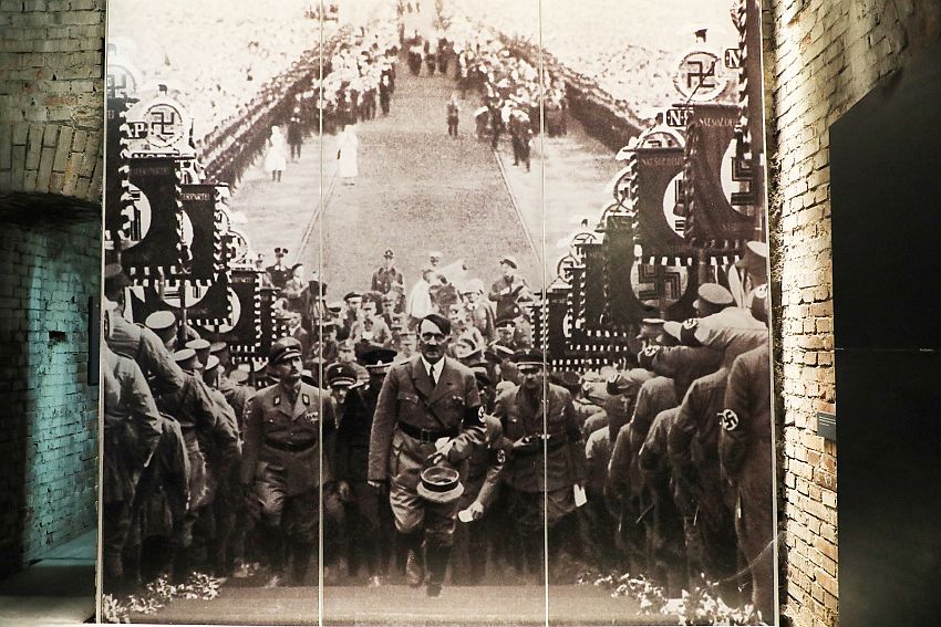 A large black and white photo fills a wall, edged on the right by a brick side wall and on the right by an arched doorway. The photo shows Hitler in uniform, climbing a stairway directly toward the viewer/photographer. Lining both sides of the stairways are soldiers, facing down the stairs,  some doing the straight-armed Nazi salute, some holding flags iwth swastikas. In the background, in the field below the stairway, more soldiers line a cleared center strip. 