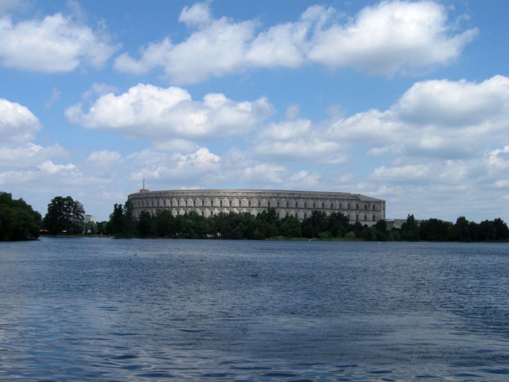 As seen across a lake, the building in the background, the Nuremberg Documentation Center, is wide and curved and several stories high, with arched windows. Blue sky with fluffy clouds above.