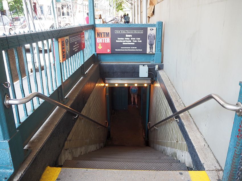 Looking down a stairway with a wall on one side and a rail on the other. A small sight on the rail straight ahead says "New York Transit Museum" and lists the museum's opening hours.