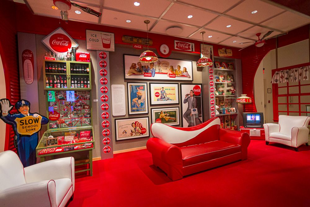 A room with two big comfortable-looking armchairs on either end, both white but edged in red, and a big sofa in the middle, bright read with a curve of white across the back. The walls of the room are covered with Coca-Cola memorabilia: vintage signs and bottles and lamps in the tradmark red and white. The floor is also bright red.