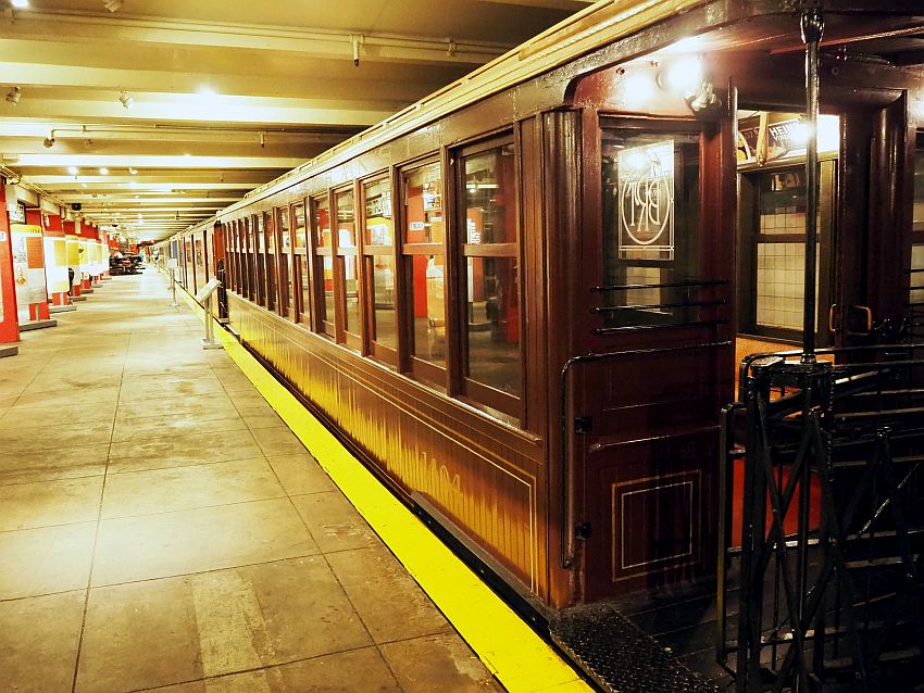 Looking down a long flat subway platform. On the right, on the tracks, is a row of subway trains. Only the nearest is really visible: it's wooden, and painted dark red.