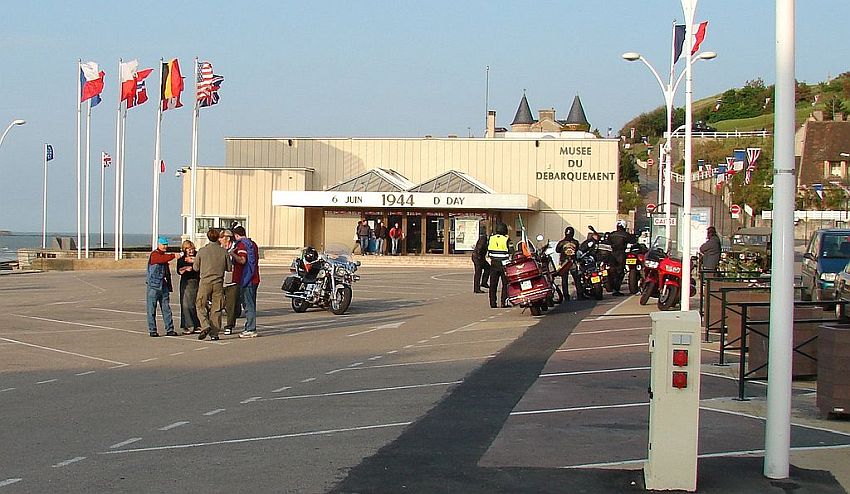 The front of the Musee du Debarquement: a simple low building without windows, off white, flat roof, with the words "6 juin 1944 D-Day" above the entrance. In the mostly empty parking lot in front are several motorcycles with their riders standing around them and talking. Beside the building are a number of flagpoles with various countries' flags flying.