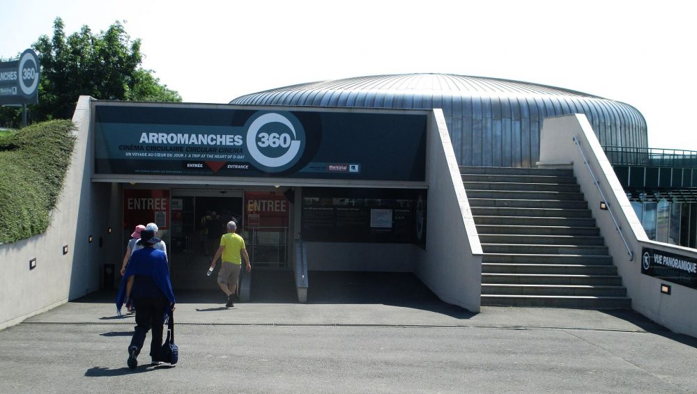 The entrance is low, and looks to be built into the side of a low hill. To the right of the entrance is a stairway up one flight to the roof of the entrance. Behind, part of the cinema itself is visible: a round low cylindrical structure. Above the entrance door: Arromanches 360.