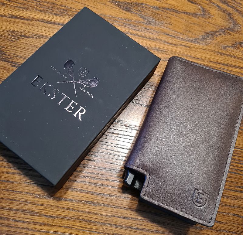 On the left, the box the wallet came in: It is black, with shiny letters that say Ekster and crossed symbols that are not very clear. On the right, a brown leather wallet, but with a black plastic knob in the left-hand corner. In the right hand corner, the letter E inside a crest shape is embossed on the leather.