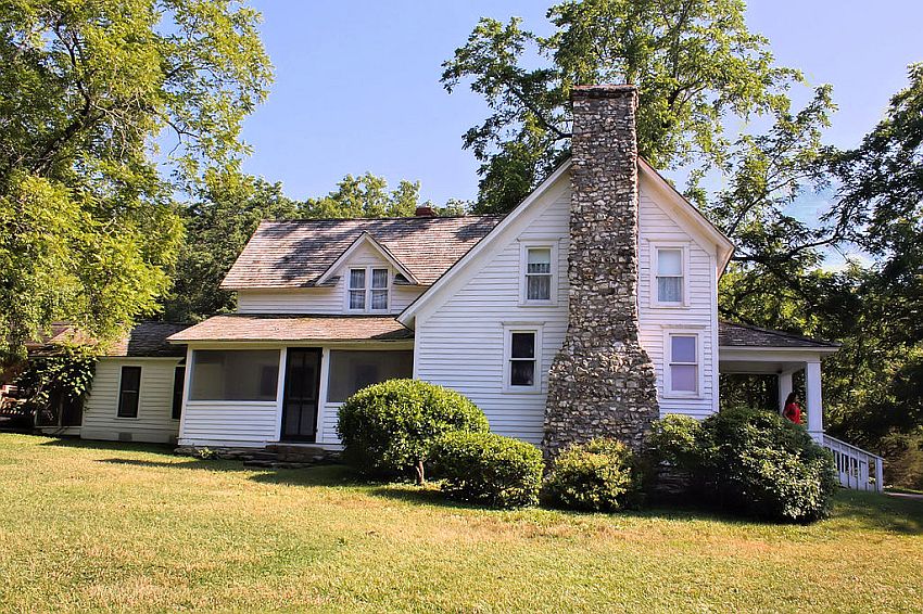 A white house in an L-shape. On the left, two stories with a closed-in porch in front and a dormer window in the center above. On the right, also 2 stories, seen from the side, with a stone fireplace going up the middle of the side. On the right-hand end, seen from the side, a veranda across the front of the house, with a short stairway going down from it.