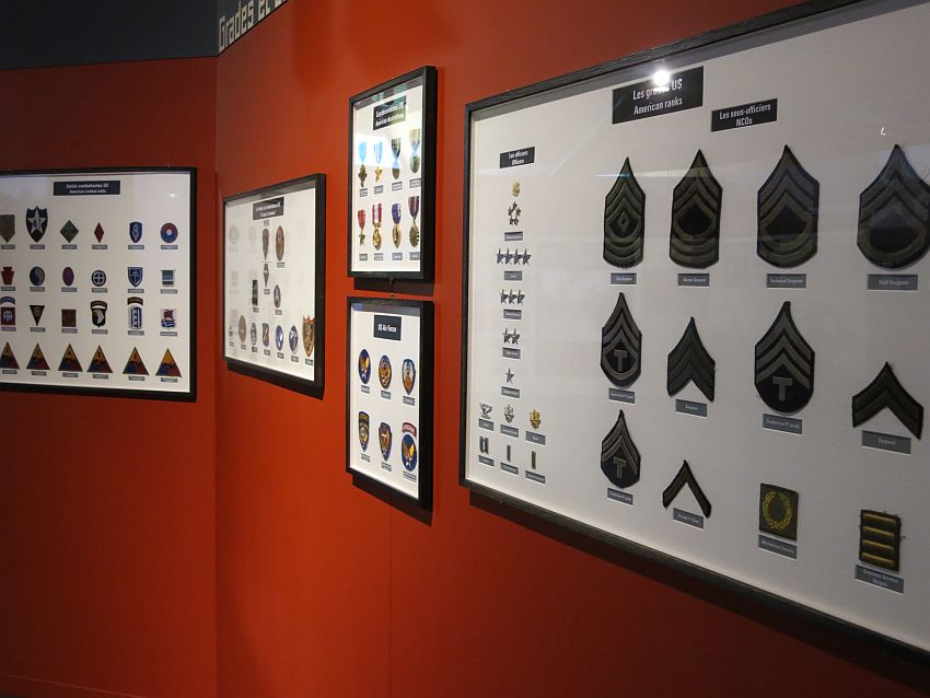 A deep red wall with a series of frames hanging on it. Each frame is filled with various insignia and medals: all things that would be attached to a uniform. The nearest one is filled with the insignia of the different American ranks in the military: shoulder patches and pins, each mounted and labeled. 