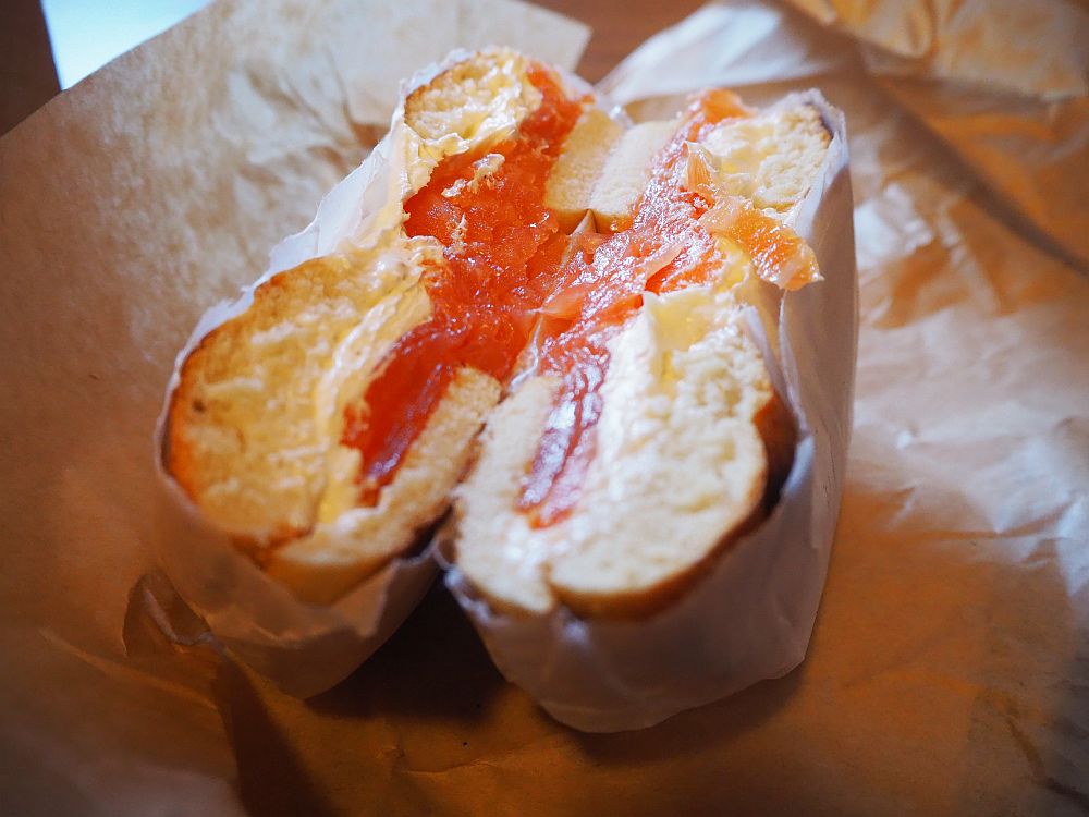 Two halves of a bagel sandwich wrapped in brown paper. The cut side faces up, showing the cream cheese and orange lox.