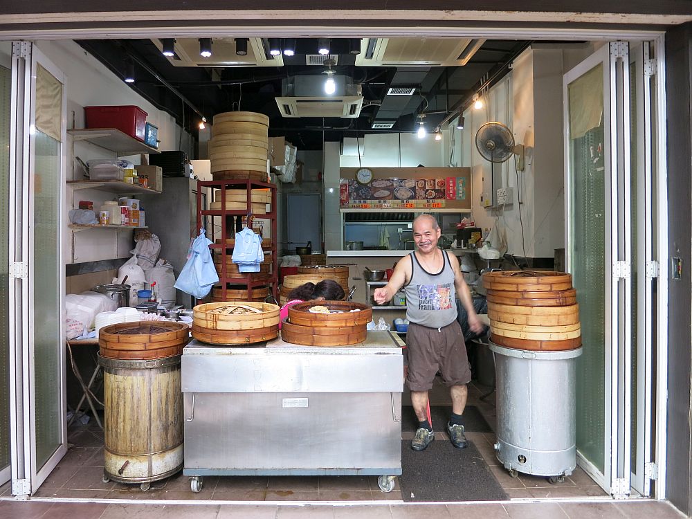 The entire storefront is open to the street with glass doors folded back to the sides. A table in the middle holds baskets used for steaming dim sum. A man, smiling in a sleeveless shirt and shorts, stands between the table and a cylindrical table of some sort with another pile of the baskets. Behind is the whole kitchen, with various supplies visible and a fan fixed to the wall. 