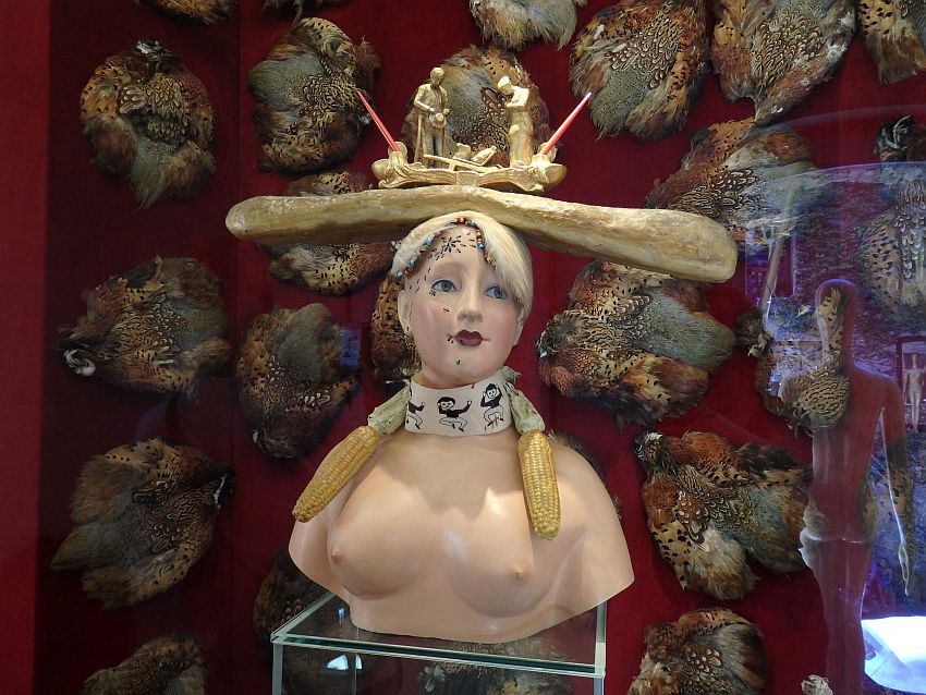 A sculpture of a woman from just under her breasts and up. She is naked, breasts exposed. Around her neck is a band with a human figure on it who appears to be jumping. An ear of corn sits on either shoulder, hanging in front like pigtails. She has ants crawling on her forehead and down to her mouth. On her head is a long horizontal loaf of bread: a baguette. On top of that is a carved scene: two men, both looking down, one holding his hat in front of him. It is not clear what they are looking at.