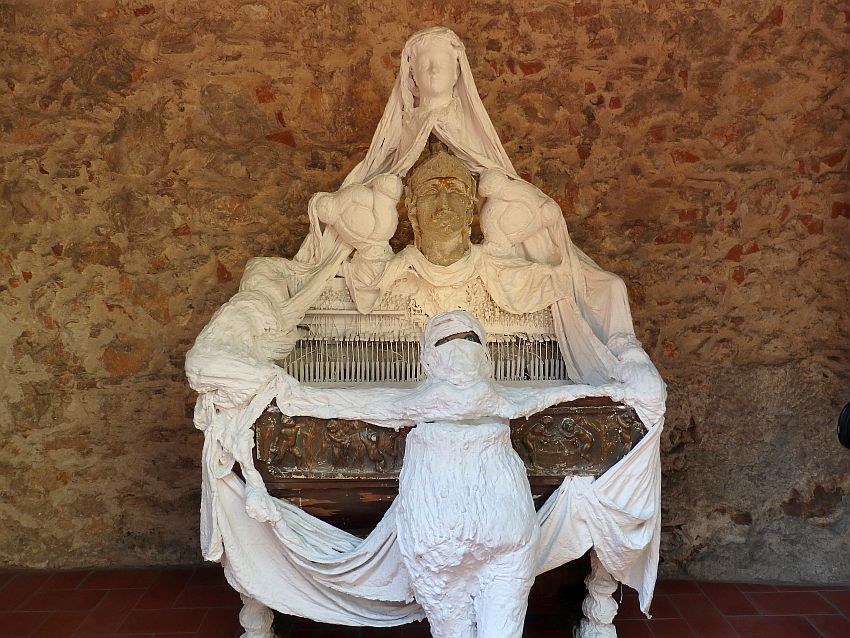Something like a harpsichord or old piano is draped with cloth and papier mache figures, all in white. In the center, on top of the piano, a persons head. Above that, another person's head. In front of the piano, a somewhat human shape made of white plaster or papier mache, with a head almost completely wrapped in except for a slit where two eyes peer out.