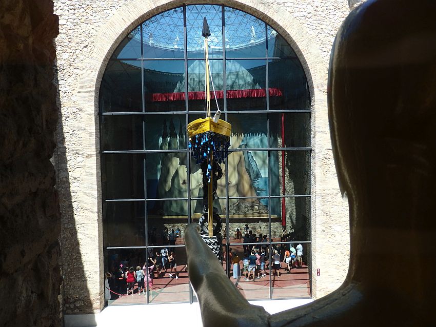 On the right is the back view of a head, with an arm sticking out straight ahead. It seems to point at the boat standing on top of a pole. Behind that is a large arched window. People are visible inside the building at the bottom of the multi-story window, mostly looking up at the huge multi-story painting on the far wall, which shows a naked torso.