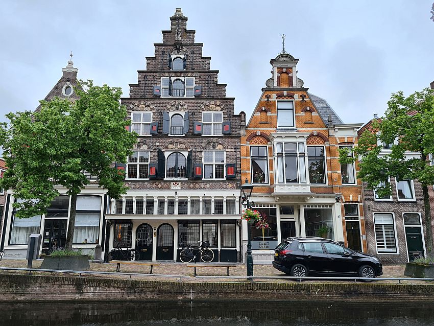 Two row houses clearly several centuries old. the left-hand one has a tall, steep step gable. The ground floor is mostly windows. Above that, two floors with 3 windows each, then 2 floors with 1 window each. On the right, a smaller house in a more orangy brick, with a bay window above the entrance.