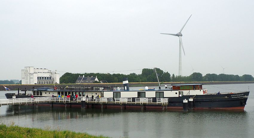 This photo shows just how long the Magnifique is, and low to the water. It is docked at a dock that has a white picket fence along its length. Beyond the ship is a very tall modern windmill.