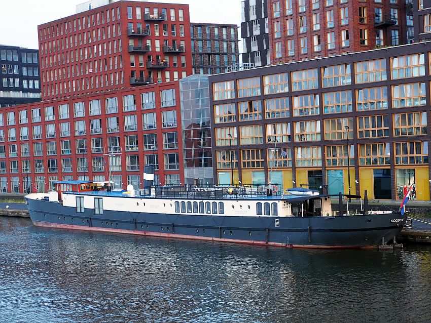 The boat is tied to the riverside and is long and low. The hull is black with a row of portholes just above the water line. Above that is painted white, and has bigger windows here and there. Behind the boat are tall buildings with lots of windows.