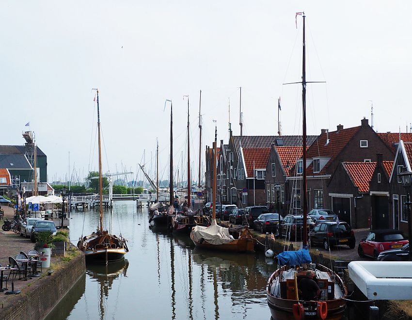 A canal with a row of houses of various shapes and sizes along the side and a row of wooden sailboats moored along the sides of the canal.