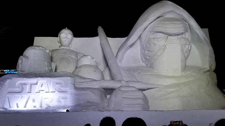 A Star Wars themed snow sculpture - it is huge in comparison with the people's heads visible at the very bottom of the photo. ON the left, R2D2 and C3PO, along with some other creature that looks like a small snowman. Below them is the Star Wars logo. To the right is a much larger figure that may be Kylo Ren in his mask and with a hood over his head.