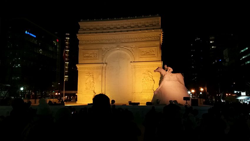 The large snow sculpture shown is lit in a yellowish light against the darkness of light. It is a large building with an arch - perhaps the Arc de Triomphe, with detailed bas-relief detail on it. To its right is a sculpture of a man riding a horse. 