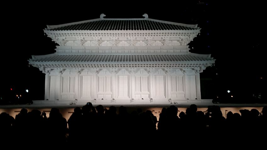 It is nighttime, but the snow sculpture at the center of the photo is well-lit. It is a detailed model of a building, perhaps a traditional temple with vertical columns and a tiled roof that curves up slightly at the outer edges. It seems similar to traditional palaces or temples in South Korea.