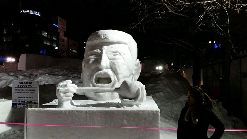 It's nighttime at the Sapporo Snow Festival, but the snow sculpture at the center of the picture is well-lit. It is a huge head that resembles Donald Trump with his mouth wide open. His hands show in front of his chin holding either end of a gavel or hammer, also all made of snow.