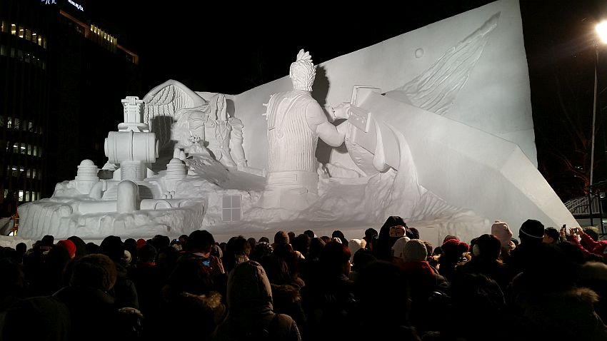 This snow sculpture at the Sapporo Snow Festival refers to some story I am not familiar with. It is absolutely huge, judging by the people who are visible at the bottom of the photo. The sculpture shows a large man onthe right with is back to the photo, perhaps with a chicken's head? He is showing from the hips up and wields a sword larger than he is by far. It appears to extend out of the sculpture into the audience. On the left is another figure further back, a human in a hooded cloak. 