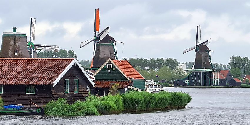 Seen across the water, three windmills, the nearest one with sails, and two low houses in front of it on the riverbank.