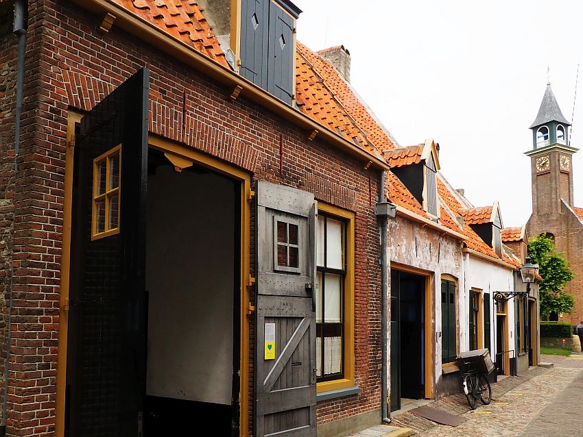 Looking down a row of building. The nearest is red brick with orange trim and large open doors that look like barn doors.The buildings behind them are houses, very small, each with a door and one window on the ground floor and one dormer window upstairs. A church tower is visible beyond them.