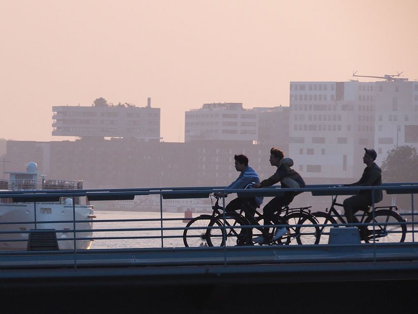 Three young men bicycle across a bridge in Amsterdam, sillhouetted against a darkening sky with tall buildings in the background.