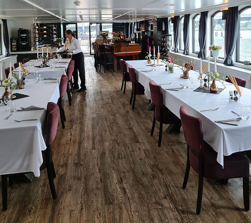 The dining room has two long tables set with white tablecloths and candlabras. the seats are set well apart from each other. A woman is busy setting the tables. 