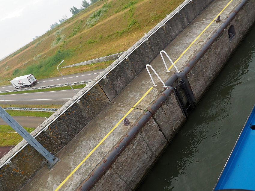 The picture is taken on the diagonal, so a bit of the boat can be seen in the bottom right corner, a strip of water beside it, then a concrete wall, then, at the top left corner, a street extends to the left, with one cargo truck visible.