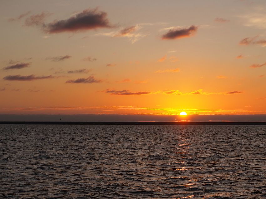 Calm water with only slight ripples. Clouds low on the horizion with half of the sun still visible. The sky is orange, with a few dark puffy clouds.