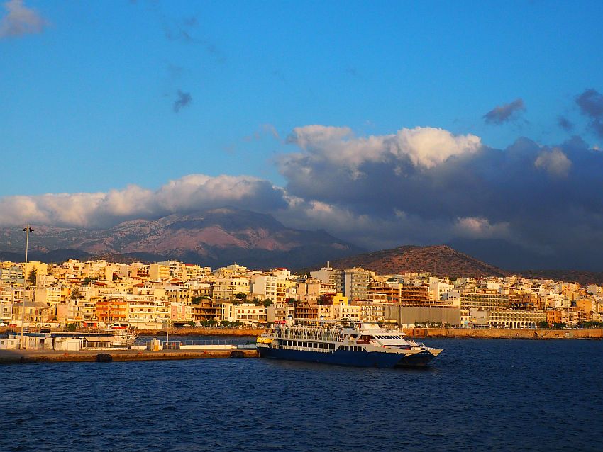 A view of the city, with many white buildings looking yellow in the low light. Beyond the city is a mountain, shrouded at its top by puffy white and gray clouds. In the foreground, the sea, and one boat is docked in the center of the picture: it looks like a small ferry.