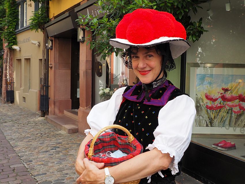 The photo shows her from the waist up. She wears a white blouse with puffy sleeves to her elbows and clutches a small basket in front of her. On top of the blouse is a vest, the top of her dress, which is black with small embroidered flowers and a wide purple-edged collar. She smiles at the camera, wearing a had with a white brim and a bunch of very large bright red pompoms on top of the hat.