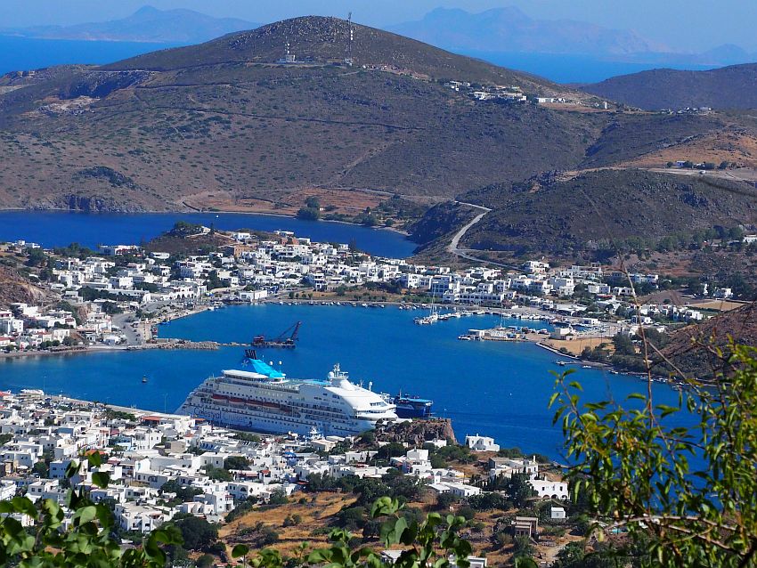 Looking down from a hill to Patmos harbor: blue water surrounded by land covered with white buildings. The ship towers over the nearest buildings. Beyond the harbor is a mountain with very few buildings on it.