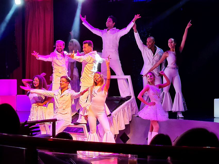 9 people - 5 men and 4 women, hold their hands up and pose, facing the audience. They are all wearing white clothing in a 70s style, e.g. exaggerated bell-bottoms.