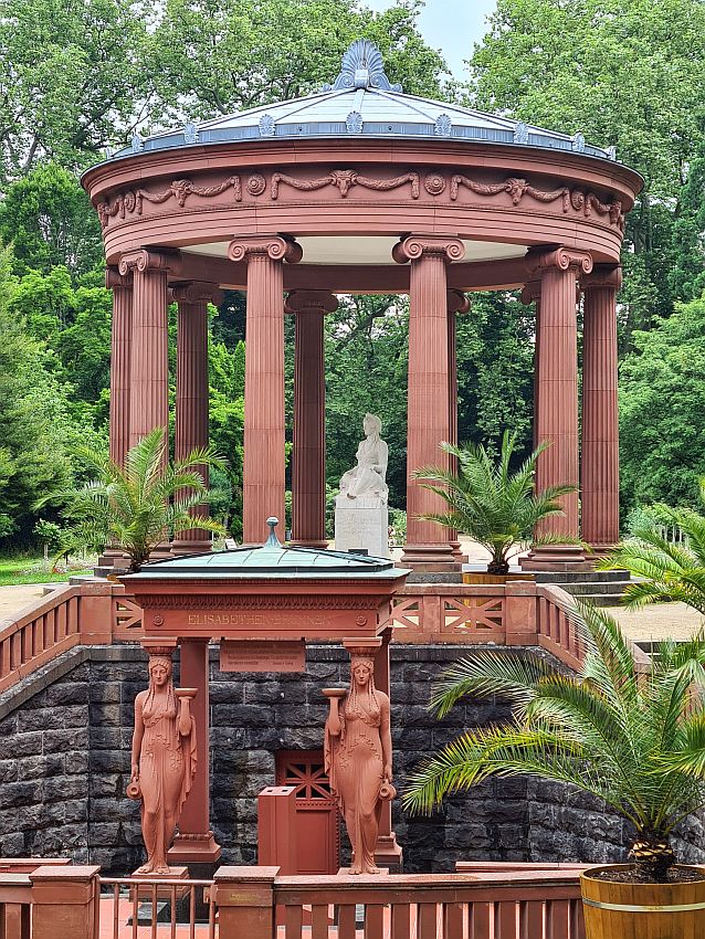 In front, a small room is held up on two classical pedestals and on the heads of two statues of women, all in dark red stone. Stairs rise on either side of this spring and above and behind the spring is a larger structure: a circle of ionic columns in the same dark red stone holds up a round roof of metal - perhaps copper. This structure shelters a statue of a woman in white stone.