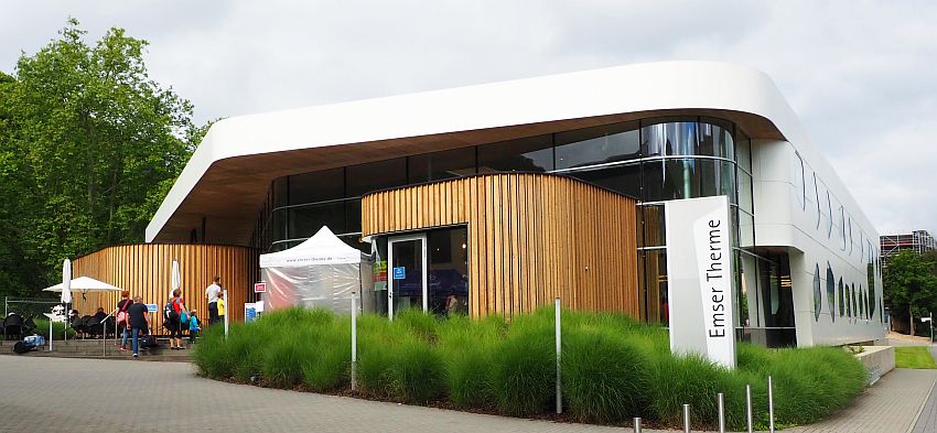 A modern building: wood slats next to the entrance, glass windows above, but the building above that and along the side is flat white. On the side the windows are round.