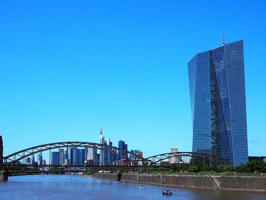 The ECB headquarters is a tall, glass-sided building at the right of the photo. It's not rectangular but leans out on one side. An iron railway bridge crosses the river at the bottom of the photo. Through the bridge the distant skyline of Frankfurt is visible: a cluster of skyscrapers.