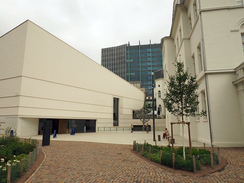 A low angular building on the left: white with only one tall window at one end and the entrance way in the center. On the right, a bit of a more traditional villa is visible, also plain white.