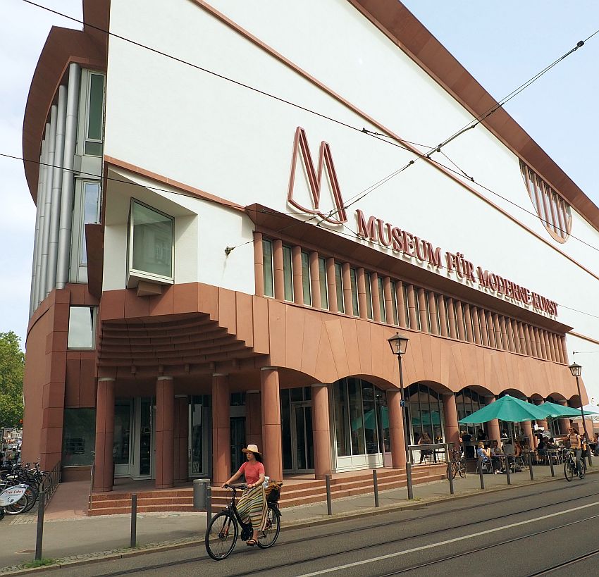 A large building on a triangular lot so the part shown has a sharp corner at the left of the photo. The building is dark red on the lower floors with arches and plain pillars and white above with no ornamentation. A cafe on the sidewalk with green umbrellas over the tables. A woman bicycles by.