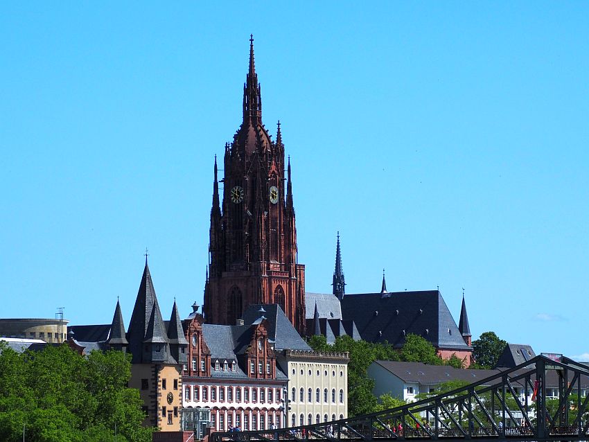 The cathedral tower is dark red stone, with pointed towers on four corners and at several heights, and with a very long narrow point on top. It has large clocks about halfway up, 2 of which are visible in this photo. In front of it, lower buildings line the river, and an iron bridge crosses the bottom of the photo.