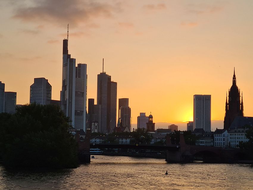 a cluster of skyscrapers seen looking along the river. Also, nearer-by, the cathedral tower, which looks as tall as some the skyscrapers in this view. All are sillhouetted against an orange sky as the sun goes down.