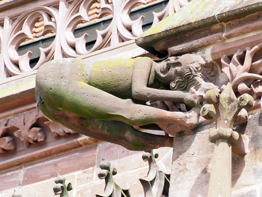 The gargoyle shown is a human figure. It is essentially folded in half, with its head, hands and feet attached to the building and its butt straight out. The drainpipe is in its butt. Its face is turned outward and it seems to be smiling.