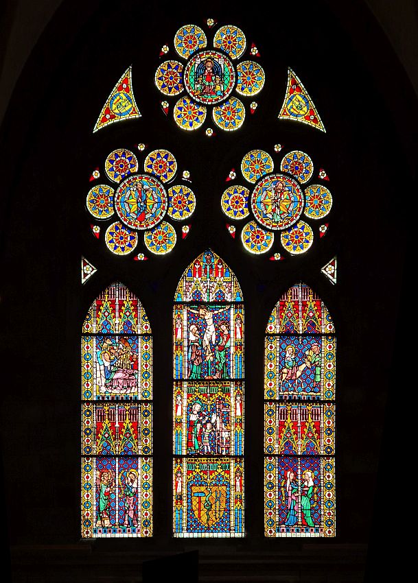 A vertical stained glass window with many many panels. At the top, 3 circular panels, below: 3 vertical panels with arched tops. The pictures visible in the glass show events in Jesus's life: the crucifixion is in the center top of the middle vertical panel. The bottom of the center panel has a cress with a sledgehammer and tongs on either side of a snake.