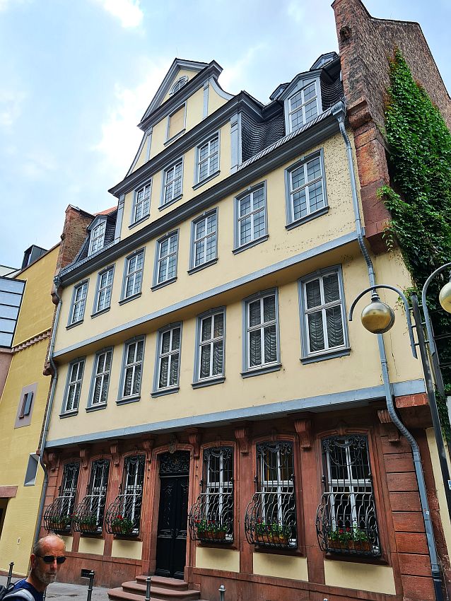 Four or five stories high, each story extends out by a foot or two beyond the story below it. It's a row house, so the sides are not visible. The ground floor is dark red stone. The floors above are plastered and painted light yellow, while the trim is grayish blue. 