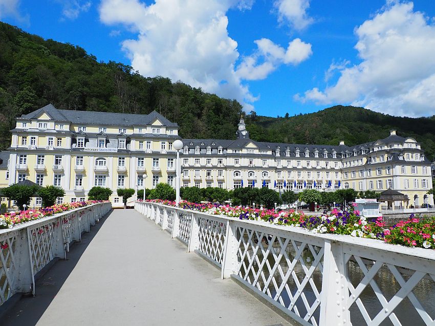 A large building of 5-6 stories and very wide: perhaps 25-30 windows across. Light yellow with white trim and many of the windows have small balconies on the upper floors. The photo is taken from a bridge across the river, and the bridge leads to the hotel and has bright flowers in boxes along both sides.