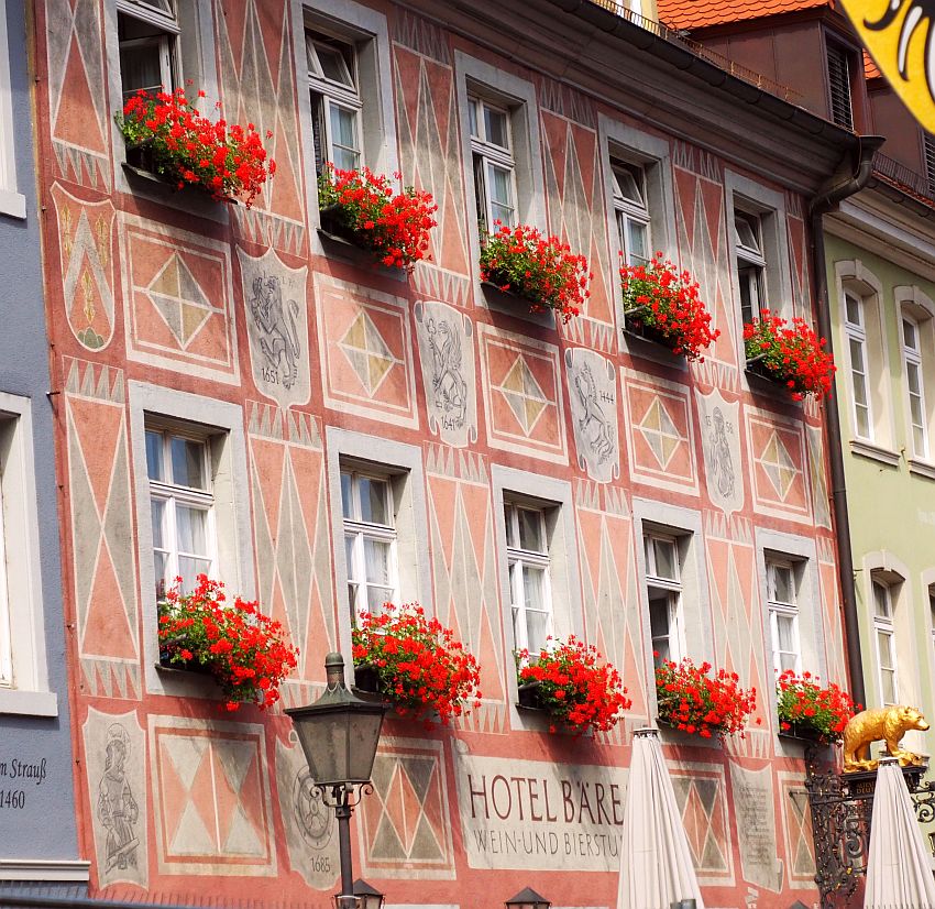 The building is flat-fronted, covered with painted plaster. The paint is pale red and light blues and is geometric patterns between the rows of windows. Between the 2 floors that are visible in this photo are images which seem to be coats of arms. Under very window - 5 above and 5 below - is a window box with bright red geraniums. 