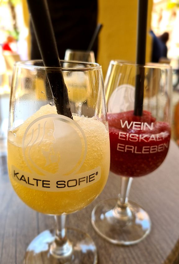 Two glasses of Kalte Sofie in wine glasses with straws. In front, the white wine version looks like a lemon slushy. Behind, the red wine version looks like a cherry slushy and says "Wein Eiskalt Erleben" which I think means "Experience ice cold wine".