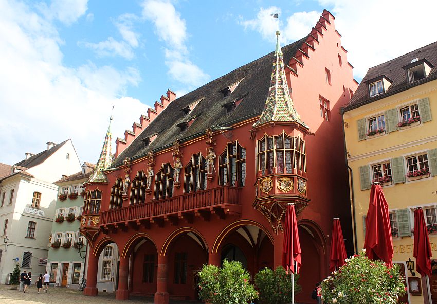 The building is painted red. It has a steep roof and step gables on either side. The ground floor has a row of pillars with arches between them, making a portico along the front of the building. The floor above that has 5 large arched windows with a narrow balcony in front of them, and at either corner of the building is a round turret with a pointed roof.